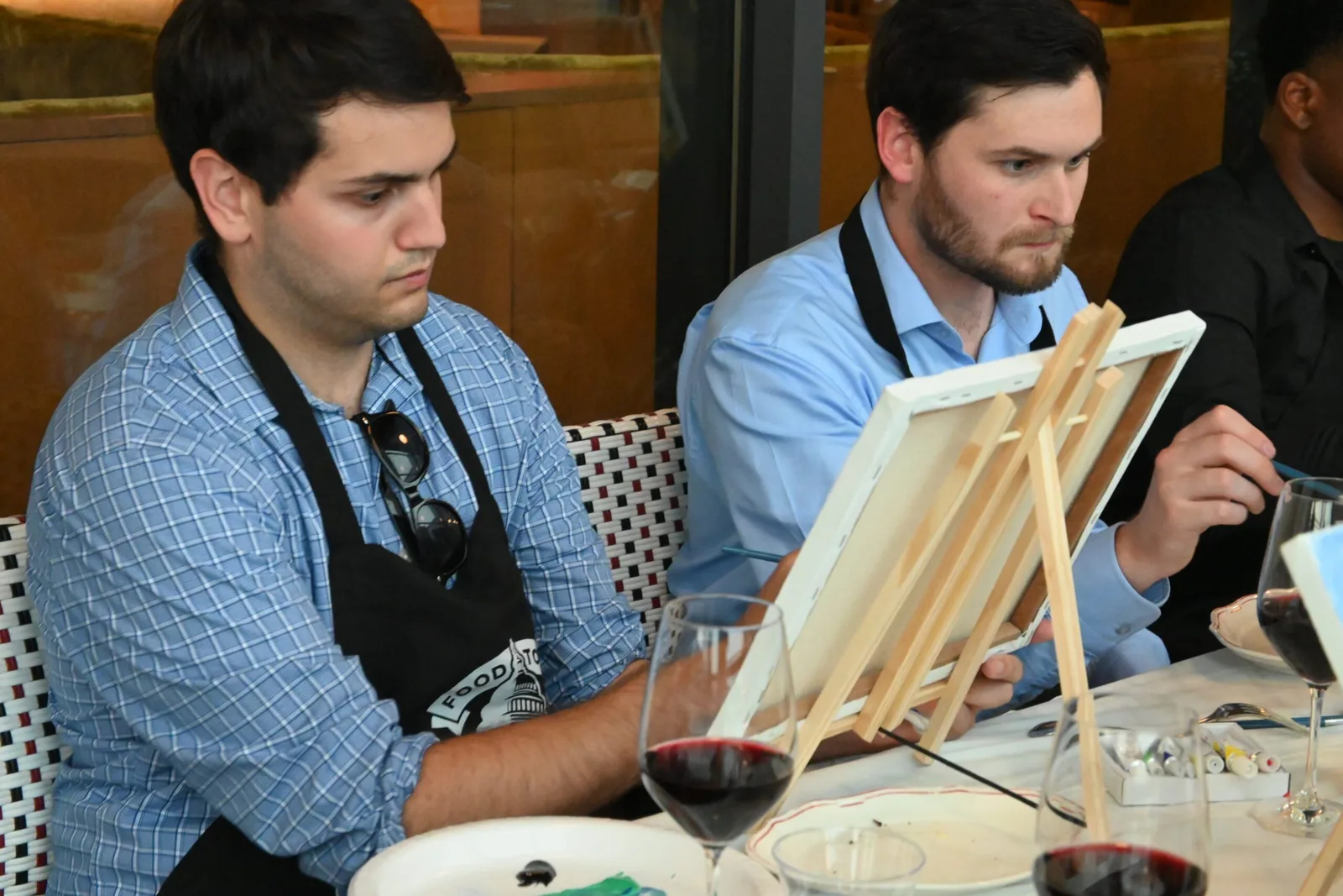 Two guests in aprons painting canvases with glasses of red wine